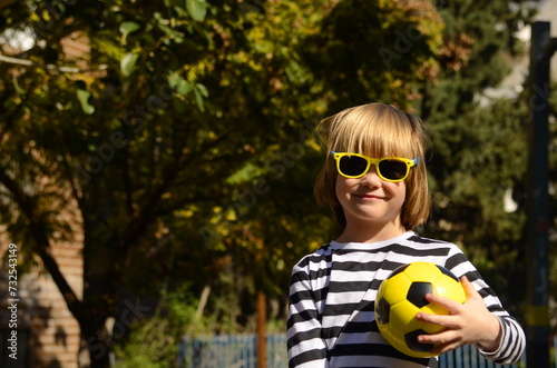 Smiling little boy, 5 years old, holding a yellow soccer ball. A child in sunglasses, a summer day, enjoys the holidays, plays in kindergarten