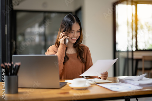 Engaged young businesswoman talking on a mobile phone while holding documents, displaying a confident smile in a bright office environment..
