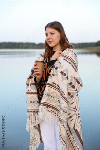 the girl is standing on a wooden pier near the lake wrapped in a blanket and drinking coffee. aesthetic photo. a glass with coffee in the hands of a girl.