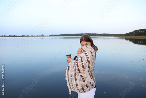 the girl is standing on a wooden pier near the lake wrapped in a blanket and drinking coffee. aesthetic photo. a glass with coffee in the hands of a girl.