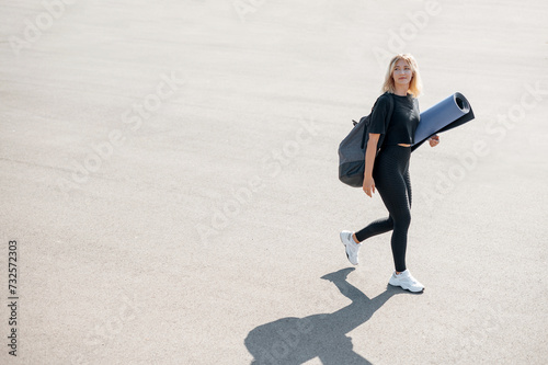 Blonde young woman walkingon the street and carrying a yoga mat while preparing for the training at the open air. Healthy lifestyle concept. Copy space