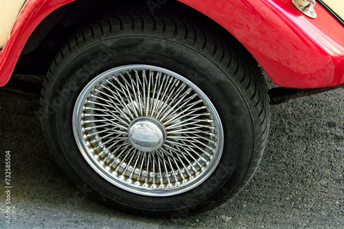 The image shows a close-up of a car’s wheel with a shiny, intricate wire-spoke hubcap. The car has a red body and is parked on asphalt.