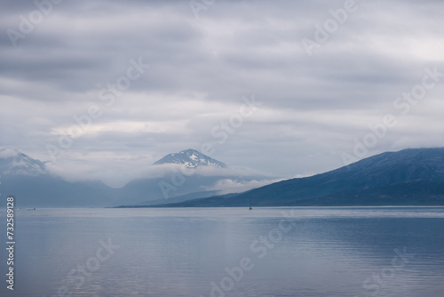 A sailboat drifts in the vastness of a Norwegian fjord, embraced by the shrouded peaks of the surrounding mist-kissed mountains