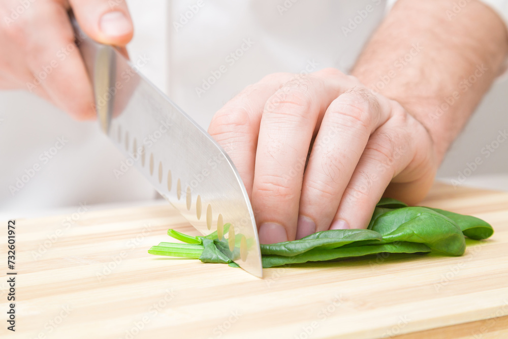Young adult man hands cutting fresh green spinach leaves with big knife on wooden board at kitchen. Closeup. Front view. Young adult man hands cutting fresh green spinach leaves with big knife on wooden board at kitchen. Closeup. Front view.