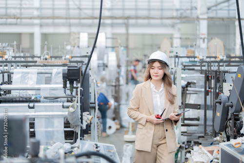 Asian female engineer walks and tests machinery in the plastic and steel industry of an export logistics company. Wearing a suit, safety helmet, laptop, radio, workers are working.