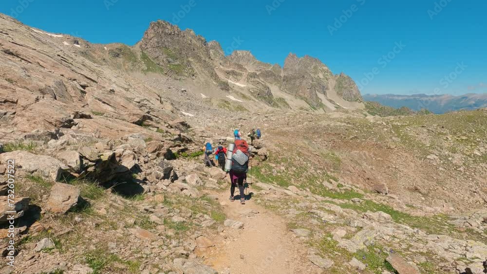 A group of tourists with backpacks walking along a trail in the mountains. First-person view of a tourist walking at the end of the group. In the background beautiful high mountains and blue sky A group of tourists with backpacks walking along a trail in the mountains. First-person view of a tourist walking at the end of the group. In the background beautiful high mountains and blue sky
