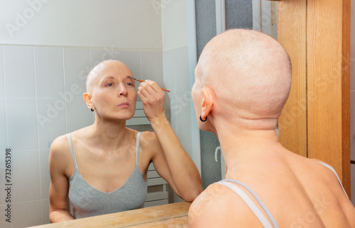 Bald woman, cancer patient applying eyebrow makeup in mirror