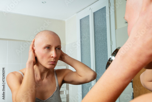 Sick woman with shaved head examining self in mirror closely