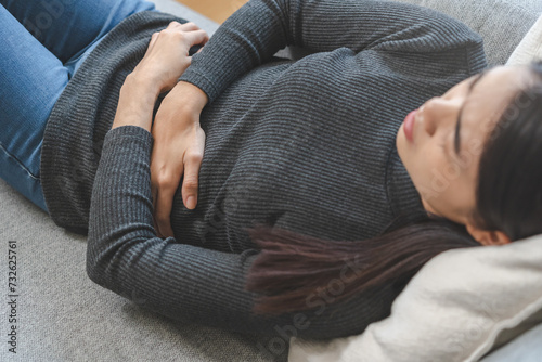 Flatulence asian young woman hand in stomach ache, suffer from food poisoning, abdominal pain and colon problem, gastritis or diarrhoea lying on sofa at home. Patient belly, abdomen or inflammation.