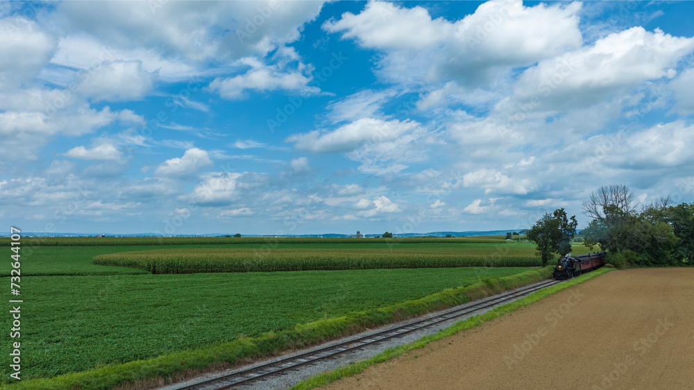 Railroad Tracks Curve Gently Through A Lush Agricultural Landscape With Fields Of Various Shades Of Green Under A Wide Sky With Broken Clouds.