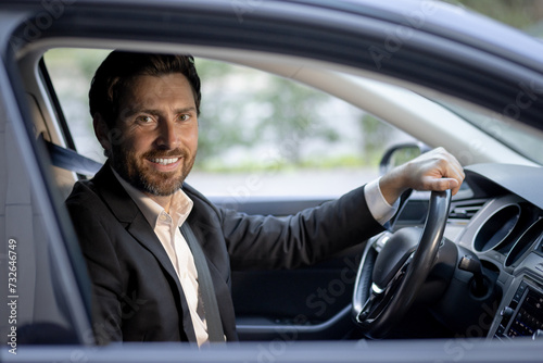 Close-up portrait of a smiling and successful young businessman sitting in the car interior behind the wheel, confidently looking at the camera