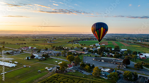 Colorful Hot Air Balloon Floating Above A Scenic Rural Landscape With Farms, Fields, And A Small Town During Sunset.