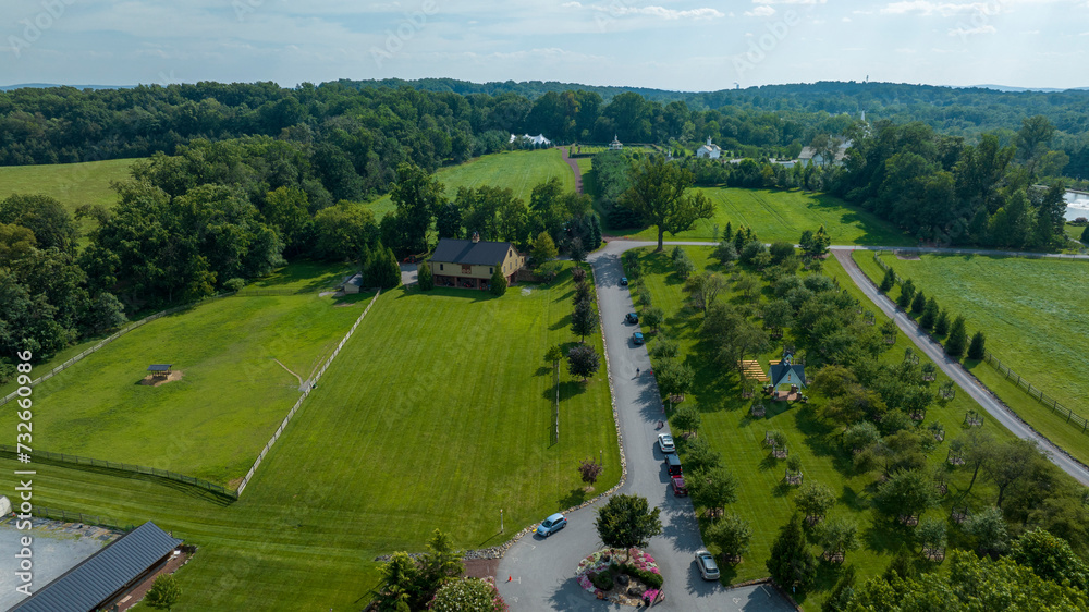 Aerial View Of A Lush Green Estate With Neatly Trimmed Lawns, A Winding Road, Fenced Paddocks, And Several Buildings Amidst A Backdrop Of Dense Trees And Far-Off White Structures Under A Clear Sky. Aerial View Of A Lush Green Estate With Neatly Trimmed Lawns, A Winding Road, Fenced Paddocks, And Several Buildings Amidst A Backdrop Of Dense Trees And Far-Off White Structures Under A Clear Sky.