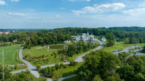 Expansive Aerial View Over A Religious Complex With Multiple White Buildings And Shining Golden Domes, A Pond With A Fountain, Set In A Verdant Landscape With A Large American Flag, With Fluffy Clouds © Greg Kelton