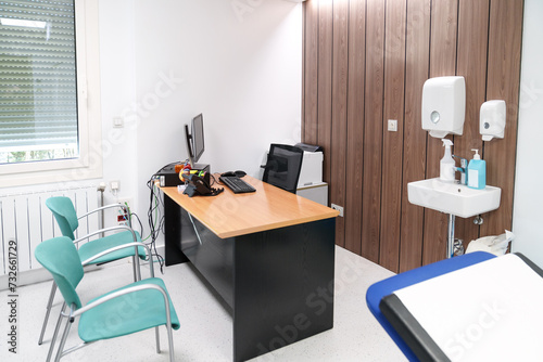 Clean and orderly doctor's consultation room with a wood desk, computer, hygiene amenities, and turquoise waiting chairs.