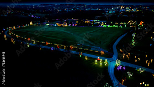 Aerial Night View Of A Winding Road Adorned With Luminous Christmas Trees Leading To A Centrally Lit Large Tree And Buildings.