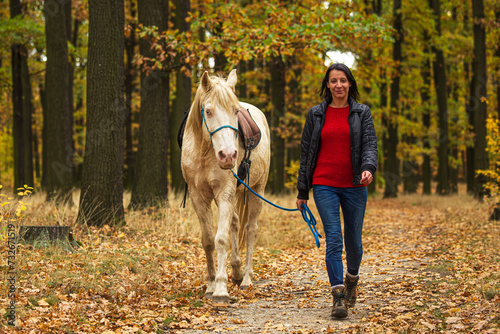 A pretty young woman and a white horse walking through the autumn colored forest © michal
