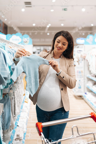Beautiful young pregnant mother choosing and buying colorful clothes and appliances for her new incoming baby in. Child shop or store concept.