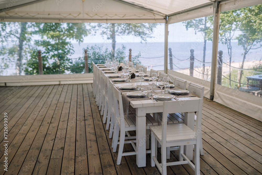 Fototapeta premium Jurmala, Latvia - july 25, 2023 - Outdoor dining setup under a tent on a wooden deck with a long table set for a meal, overlooking a beach and the sea.