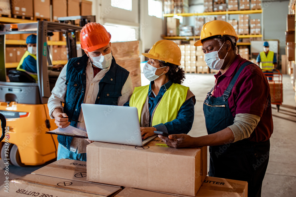 Warehouse workers in safety gear discussing logistics with laptop Warehouse workers in safety gear discussing logistics with laptop