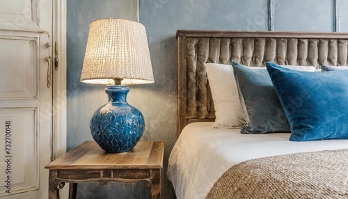 close up of blue ceramic lamp on nightstand near bed with beige fabric headboard and blue pillows and blanket french country provence interior design of modern bedroom