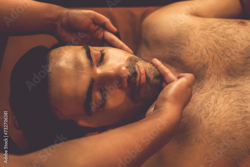 Caucasian man enjoying relaxing anti-stress head massage and pampering facial beauty skin recreation leisure in warm candle lighting ambient salon spa in luxury resort or hotel. Quiescent