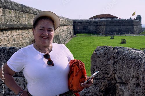 Portrait of an older woman from the waist up on the walls of Cartagena, dressed in a white blouse, wearing a visor on her head, and holding an orange Wayuu backpack and her phone in her left hand.