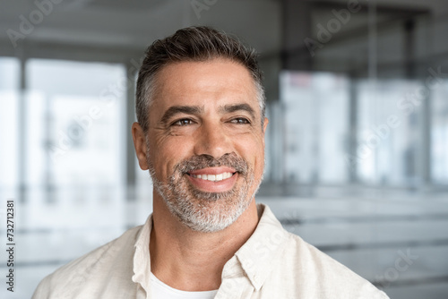 Headshot close up portrait of indian or latin confident mature good looking middle age leader, ceo male businessman on blur office background. Handsome hispanic senior business man smiling aside.