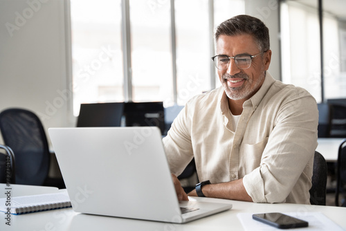 Happy smiling mature Indian or Latin business man ceo trader using computer, typing, working in modern office, doing online data market analysis, thinking planning tech strategy looking at laptop.