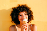 Portrait of a smiling curly afroamerican woman by a yellow wall with eating a icecream cone,