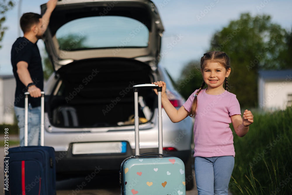 Dad and daughter are standing with their suitcases near a car with an open trunk. Happy little girl preparing to travel.