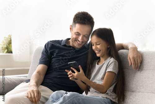 Cheerful pretty daughter kid showing funny online content on cell to happy daddy. Positive caring father and little schoolkid girl using Internet technology on mobile phone together