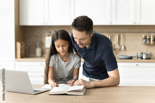 Positive caring young dad helping daughter schoolkid with homework task in home kitchen. Pre teen pupil girl and father reading book, writing essay at laptop together, enjoying family teamwork