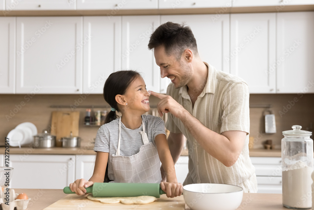 Cheerful baker father touching face of adorable daughter girl in apron with floury finger, having fun, laughing, rolling dough on home kitchen table, baking homemade bakery food Cheerful baker father touching face of adorable daughter girl in apron with floury finger, having fun, laughing, rolling dough on home kitchen table, baking homemade bakery food