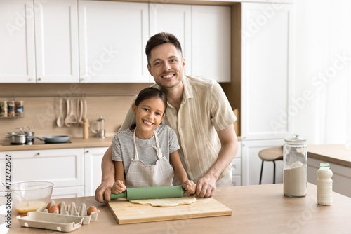 Cheerful preteen kid girl and happy young dad baking pastry food in home kitchen, rolling raw fresh dough on table, together, enjoying culinary hobby, looking at camera, smiling
