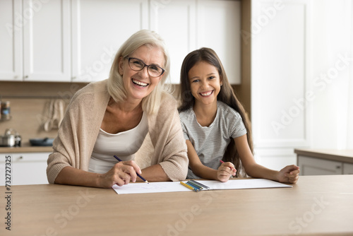 Happy pretty blonde grandma and adorable preteen grandkid girl drawing together at home, sitting at table with paper and colorful pencils, looking at camera, laughing , smiling for portrait