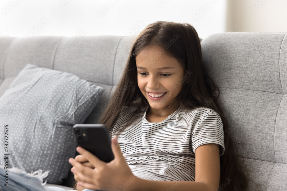 Happy beautiful pre teen girl kid browsing social media on smartphone, chatting online, playing virtual game on Internet, resting on sofa, enjoying leisure, online communication Happy beautiful pre teen girl kid browsing social media on smartphone, chatting online, playing virtual game on Internet, resting on sofa, enjoying leisure, online communication