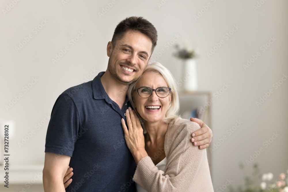 Positive handsome son man and happy blonde mature mother hugging at home with love, looking at camera with toothy smiles, laughing, standing close for family portrait Positive handsome son man and happy blonde mature mother hugging at home with love, looking at camera with toothy smiles, laughing, standing close for family portrait