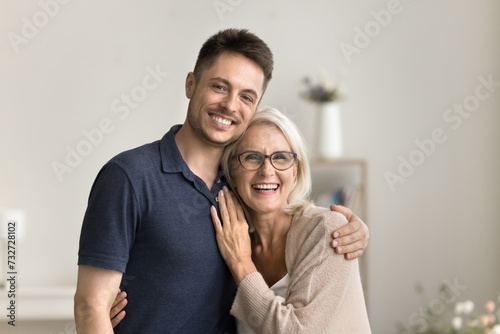 Positive handsome son man and happy blonde mature mother hugging at home with love, looking at camera with toothy smiles, laughing, standing close for family portrait