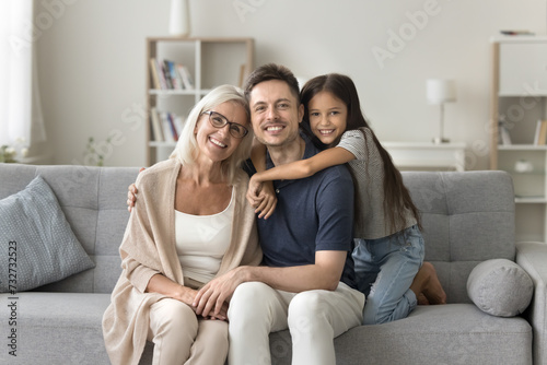 Happy pretty blonde grandmother, father and granddaughter kid hugging with love, care, affection, enjoying close relationship, family leisure at home, looking at camera, smiling for portrait