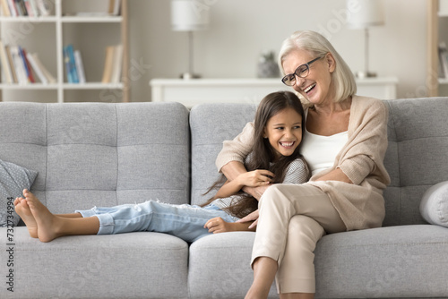 Cheerful grandma and happy pretty granddaughter kid resting on home sofa together, talking with trust, laughing, hugging with love, care, having fun, enjoying leisure, family weekend