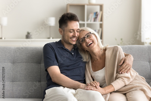 Cheerful mature mother and adult child man sitting together on couch, hugging with warmth, tenderness, love, laughing, talking, enjoying family meeting, friendship, good relationship, motherhood