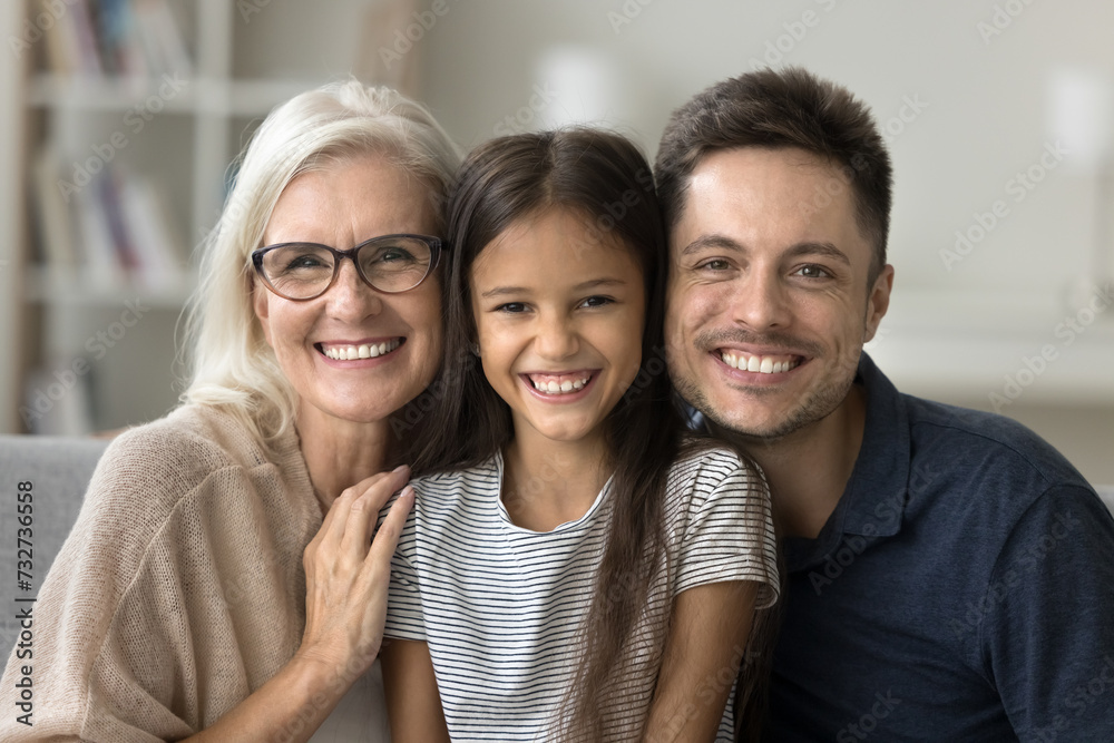 Happy cute little girl, young dad and grandmother sitting close on sofa, hugging, looking at camera, laughing, enjoying family leisure at home. Daughter kid, father and grandma head shot portrait Happy cute little girl, young dad and grandmother sitting close on sofa, hugging, looking at camera, laughing, enjoying family leisure at home. Daughter kid, father and grandma head shot portrait