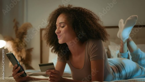 Pretty African American ethnic girl customer shopper paying credit card for internet store payment mobile phone ordering at evening home laughing young 20s woman online shopping sucessful transaction photo