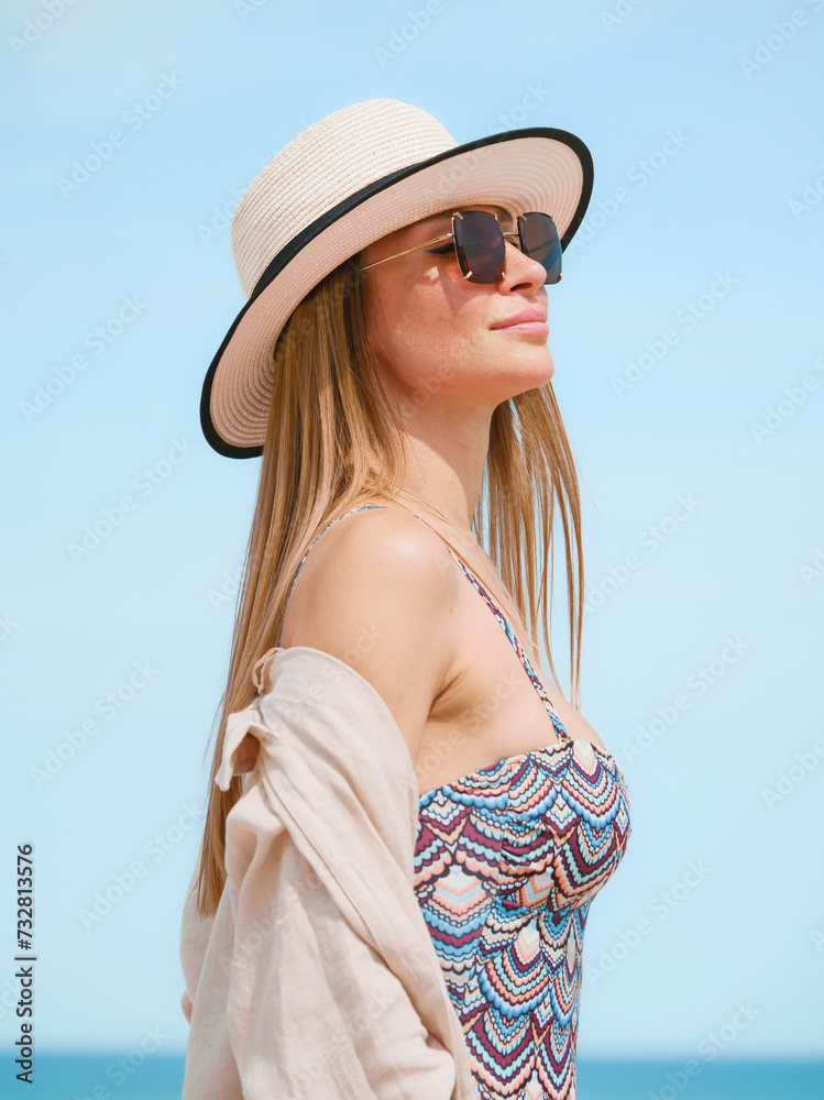 Young woman in a swimsuit, shirt, sunglasses and hat posing gracefully on a sandy beach against a blue sky
