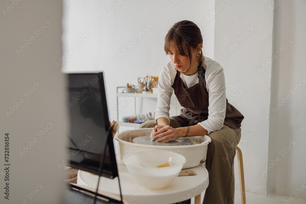 Concentrated female artisan in apron sitting on bench with pottery wheel and making clay pot Concentrated female artisan in apron sitting on bench with pottery wheel and making clay pot
