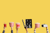 Many hands with buckets of popcorn, movie clapper and VR glasses on yellow background