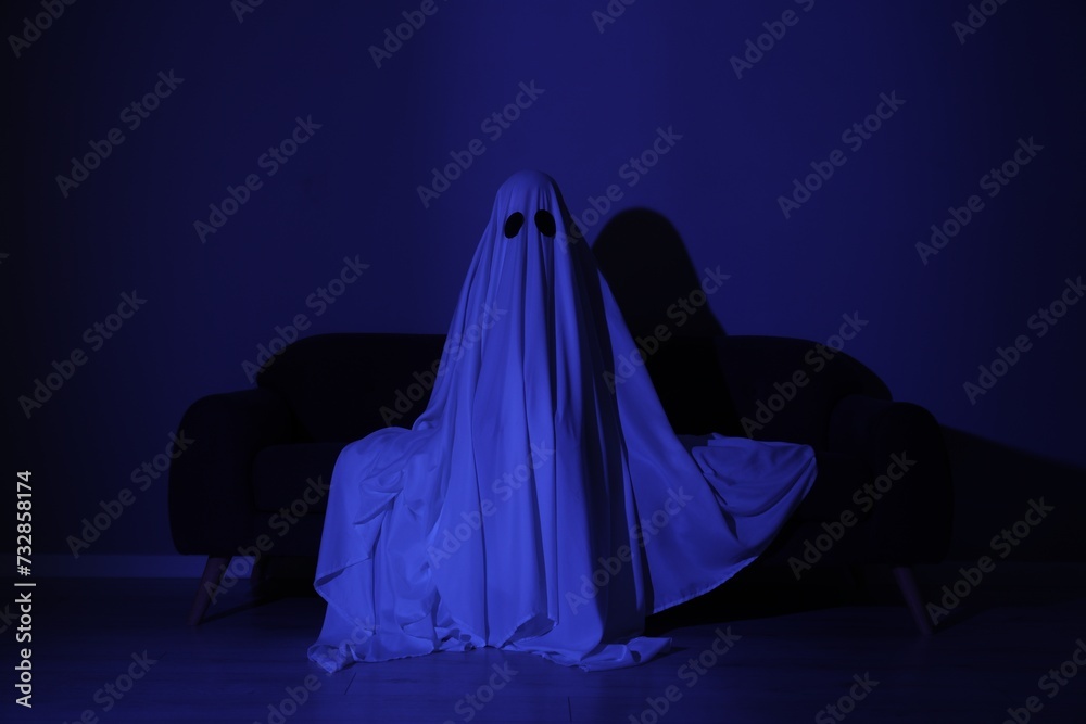 Creepy ghost. Woman covered with sheet sitting on sofa in blue light Creepy ghost. Woman covered with sheet sitting on sofa in blue light