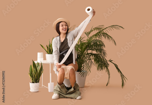 Young Asian traveler with paper roll sitting on toilet bowl against beige background photo