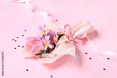 Mini bouquet of beautiful spring flowers in wrapping paper with ribbon and confetti on pink background. International Women's Day